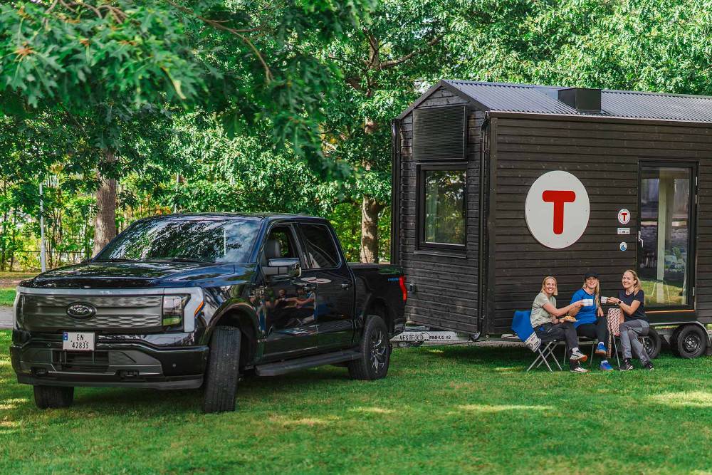 F-150 Lightning parkert ved den portable DNT-turisthytten og tre kvinner sitter utenfor og drikker kaffe.