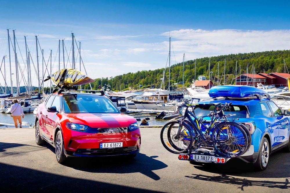 Rød helelektrisk Ford Capri med kajakk på taket og blå helelektrisk Ford Explorer med sykler på hengerfestestativ. 