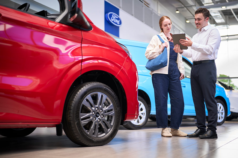 To personer i et Ford-showroom ser på et nettbrett, med en rød Ford-varebil i forgrunnen og en blå Ford-varebil bak.
