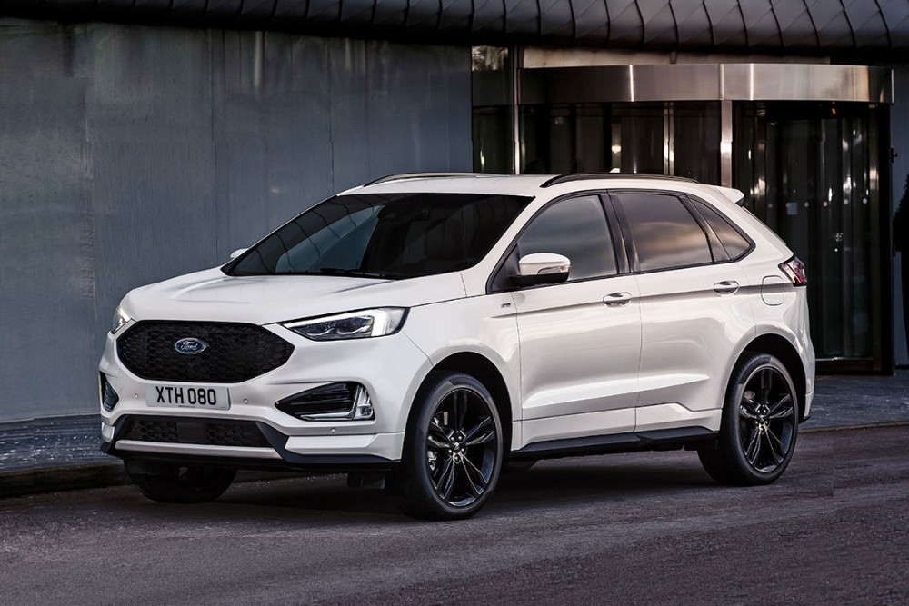 En hvit Ford Edge parkert utenfor en bygning med glassdører, sett fra front i trekvartsvinkel.