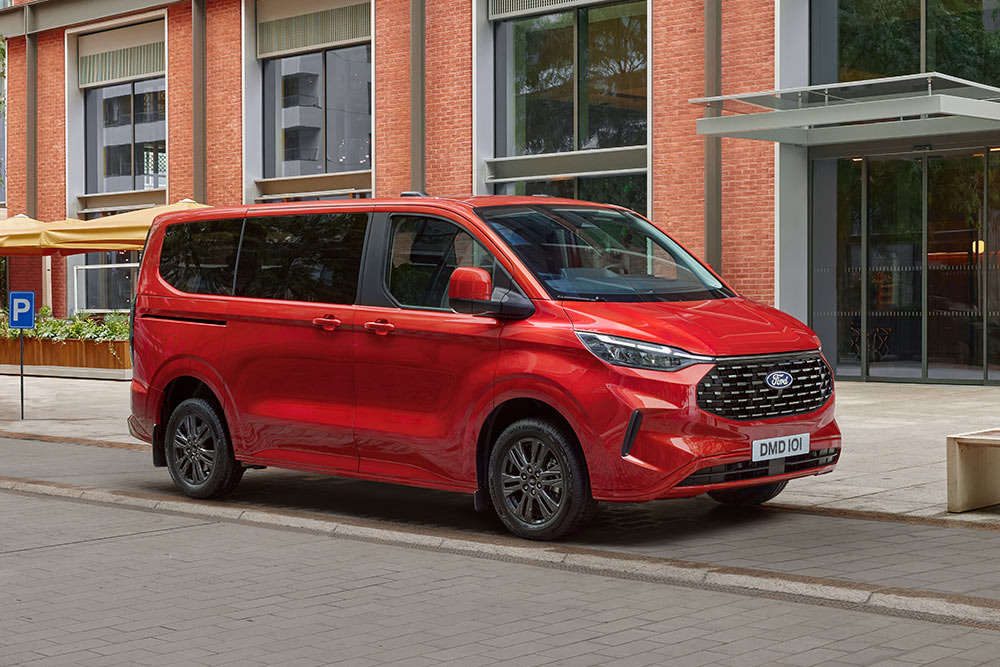 En rød Ford Tourneo Custom står parkert i en bygate foran et murhus.