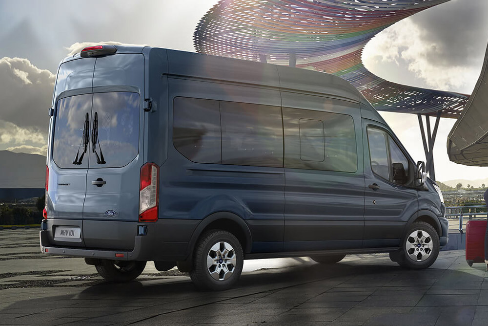 Sett skrått bakfra av en blå Ford Transit Minibuss parkert utendørs, med en moderne, fargerik konstruksjon i bakgrunnen.