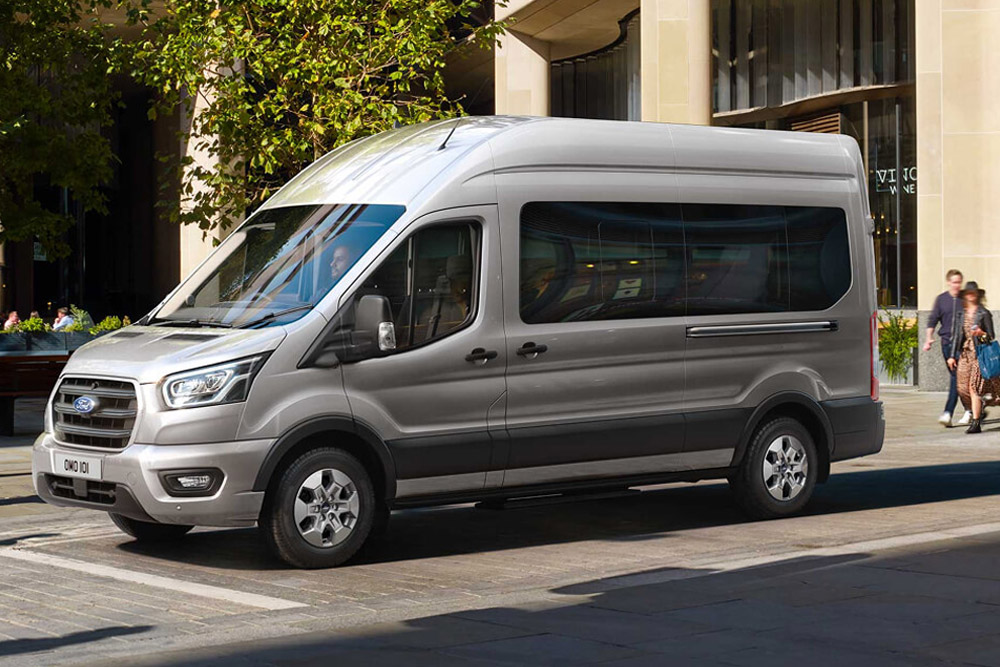 Sett skrått fra siden av en sølvfarget Ford Transit Minibuss parkert på en gate i byen, med gående mennesker i bakgrunnen.