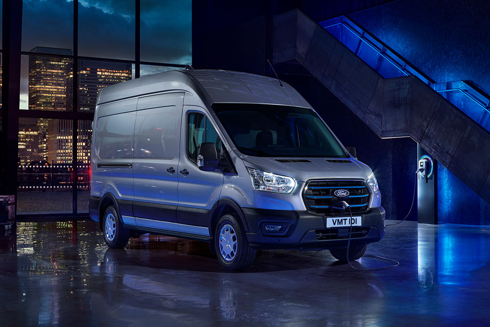 Sølvfarget Ford E-Transit elektrisk varebil parkert innendørs med en bysilhuett synlig gjennom store vinduer.