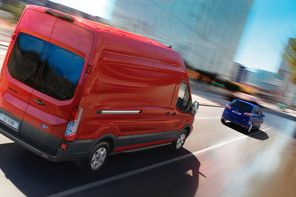Sett skrått bakfra av en rød Ford Transit som kjører på en byvei, med en blå bil foran.