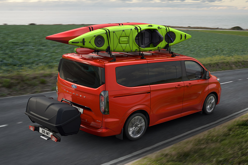 Rød Ford Pro Transit med to kajakker på taket, trekker en oppbevaringshenger, og kjører langs en kystvei.