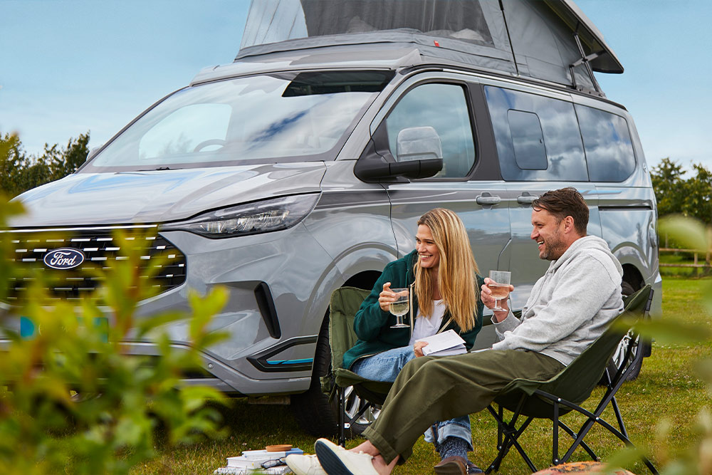Et par nyter drinker i campingstoler ved siden av en grå Ford Transit Custom bobil med et pop-up tak.