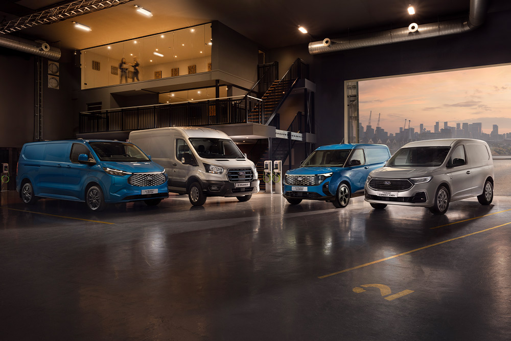 Flåte av Ford Transits i et utstillingslokale med en bysilhuett i skumringen i bakgrunnen.