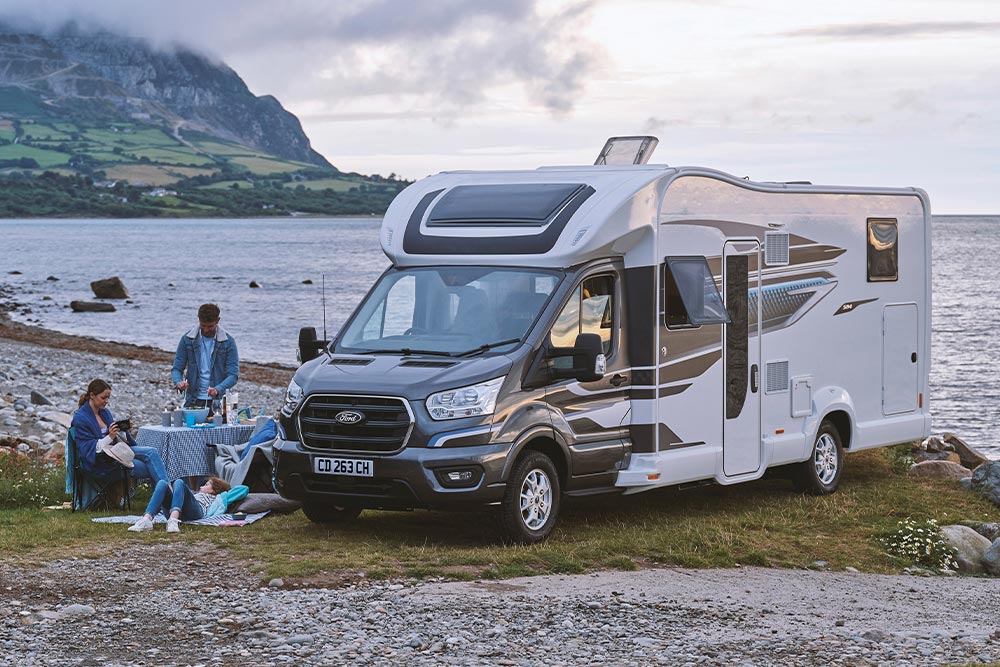 Et par nyter drinker i campingstoler ved siden av en grå Ford Transit Custom bobil med et pop-up tak.