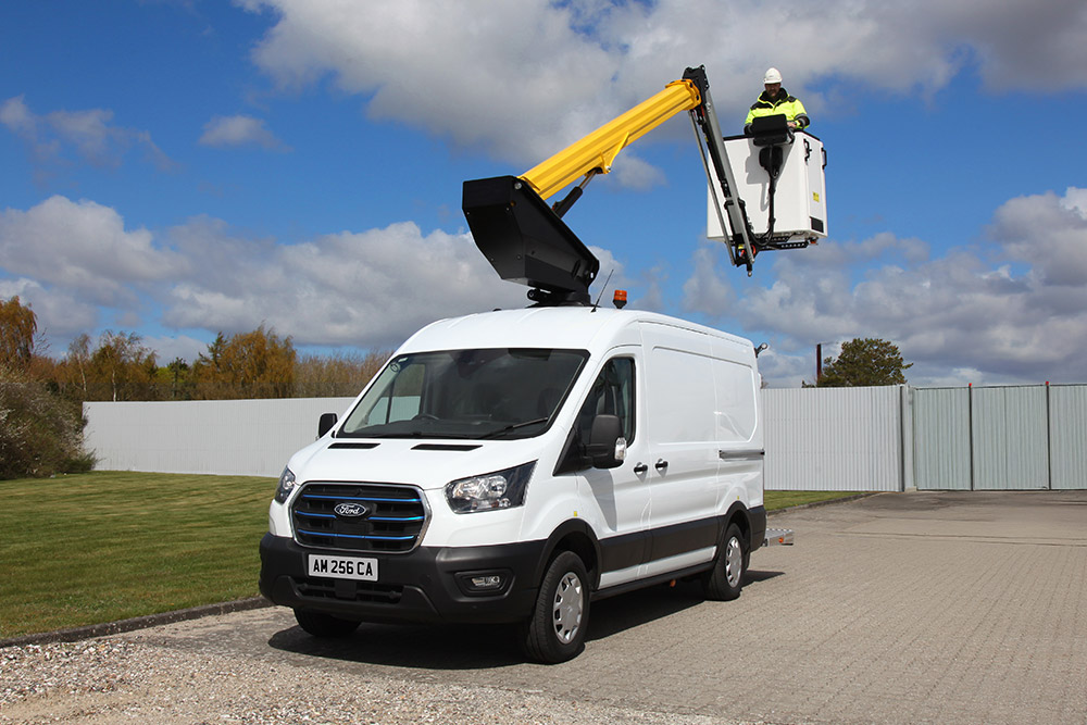 En hvit Ford Transit varebil med en utvidet lift, en tekniker i kurven, under en blå himmel.