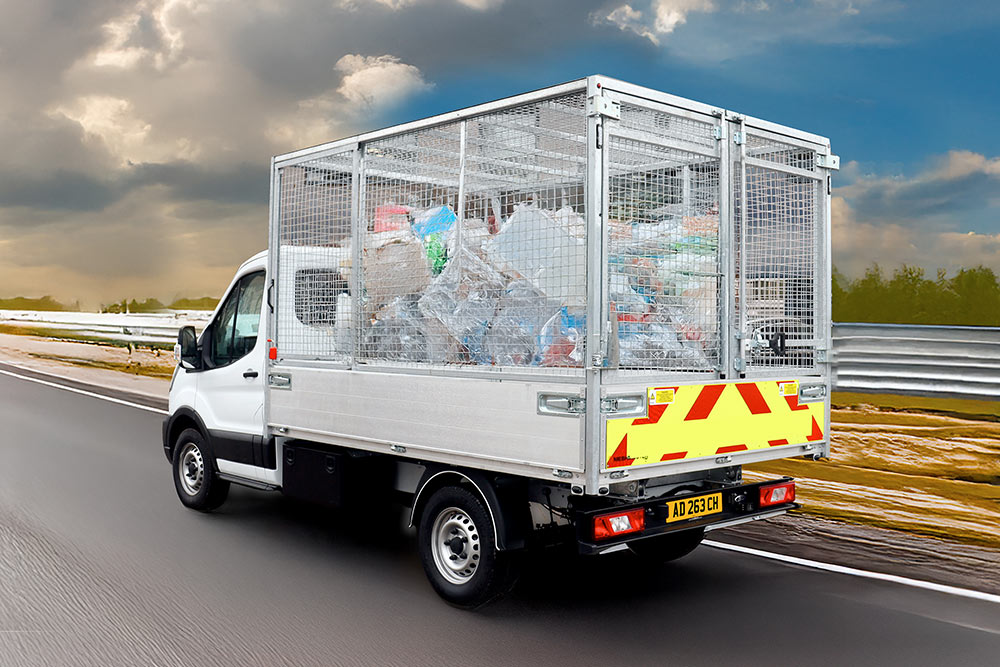 Bakfra-visning av en hvit Ford Transit tippbil med bur, full av søppel, kjører på en vei.