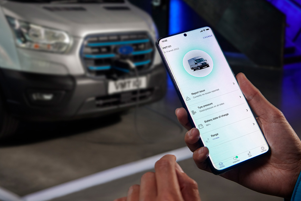 En person holder en smarttelefon som viser et app-grensesnitt, med en sølvfarget Ford Pro Transit varebil uskarpt i bakgrunnen.
