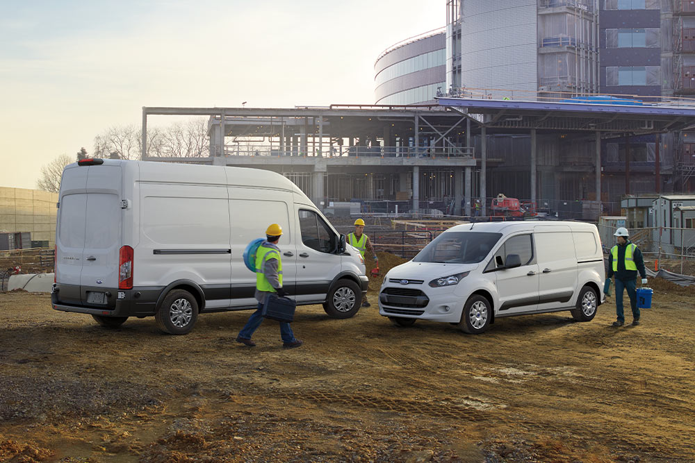 To hvite Ford varebiler parkert på en byggeplass med arbeidere som går rundt.